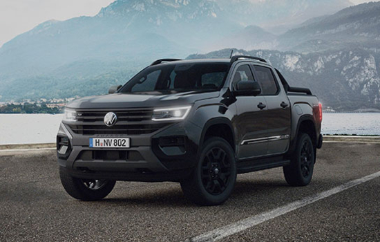 Ein dunkelgrauer Pickup mit Doppelkabine steht auf einer asphaltierten Fläche vor einer See- und Berglandschaft. Das Fahrzeug hat schwarze Felgen, markante Scheinwerfer und einen Kühlergrill mit einem runden Emblem. Die Fenster sind abgedunkelt. Im Hintergrund sind Wasser, Bäume und bewaldete Berghänge zu sehen, die von Nebel umgeben sind.